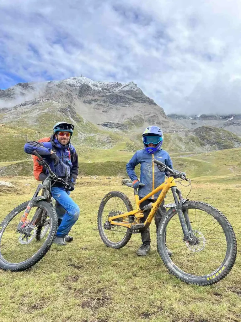 two muddied mountain bikers - a guide and an eight-year-old - smile to camera on a cloudy day in the mountains