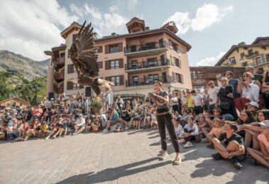 A summer crowd watches a live bird of prey demonstration in Arc 1950, with a large eagle flying close to the camera and traditional Alpine buildings in the background.