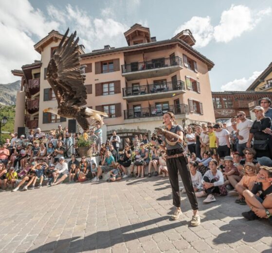 A summer crowd watches a live bird of prey demonstration in Arc 1950, with a large eagle flying close to the camera and traditional Alpine buildings in the background.