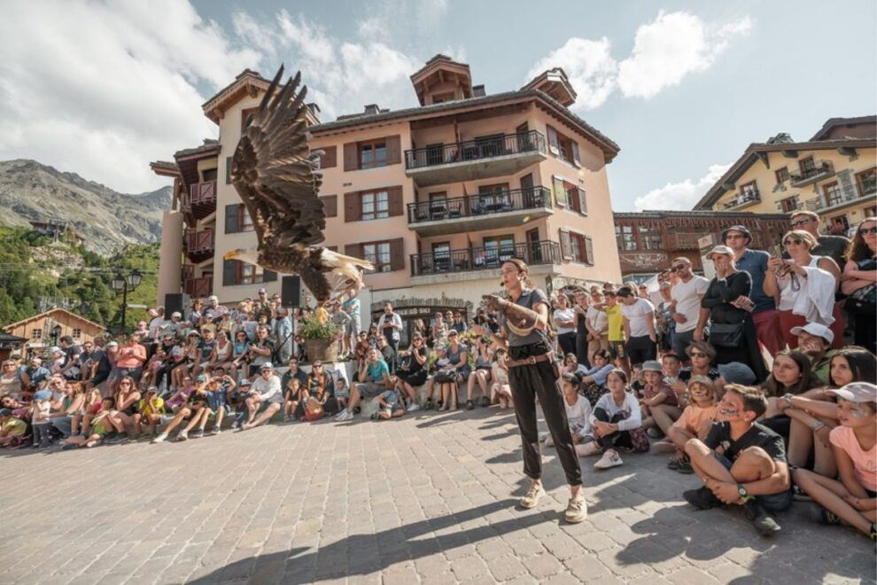 A summer crowd watches a live bird of prey demonstration in Arc 1950, with a large eagle flying close to the camera and traditional Alpine buildings in the background.