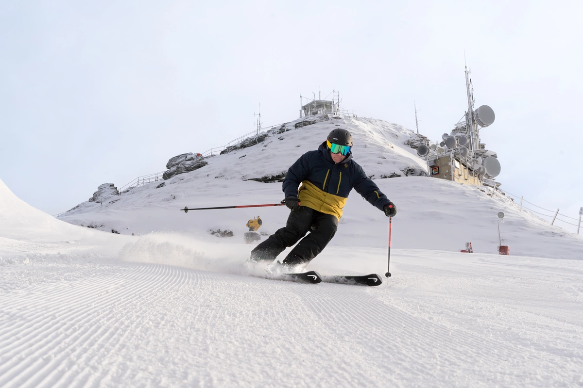 a skier on groomed piste making a turn, the peak of the ski resort's mountain behind, uphill