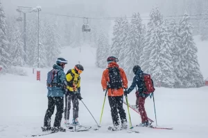 four skiers stand chatting in the snowiest of environments, under a gondola. The huge pine trees surrounding them on the piste are laden with snow