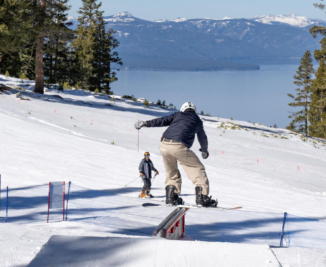 a snowboarder grinds a rail, a lake ahead in the distance with snow-topped mountains