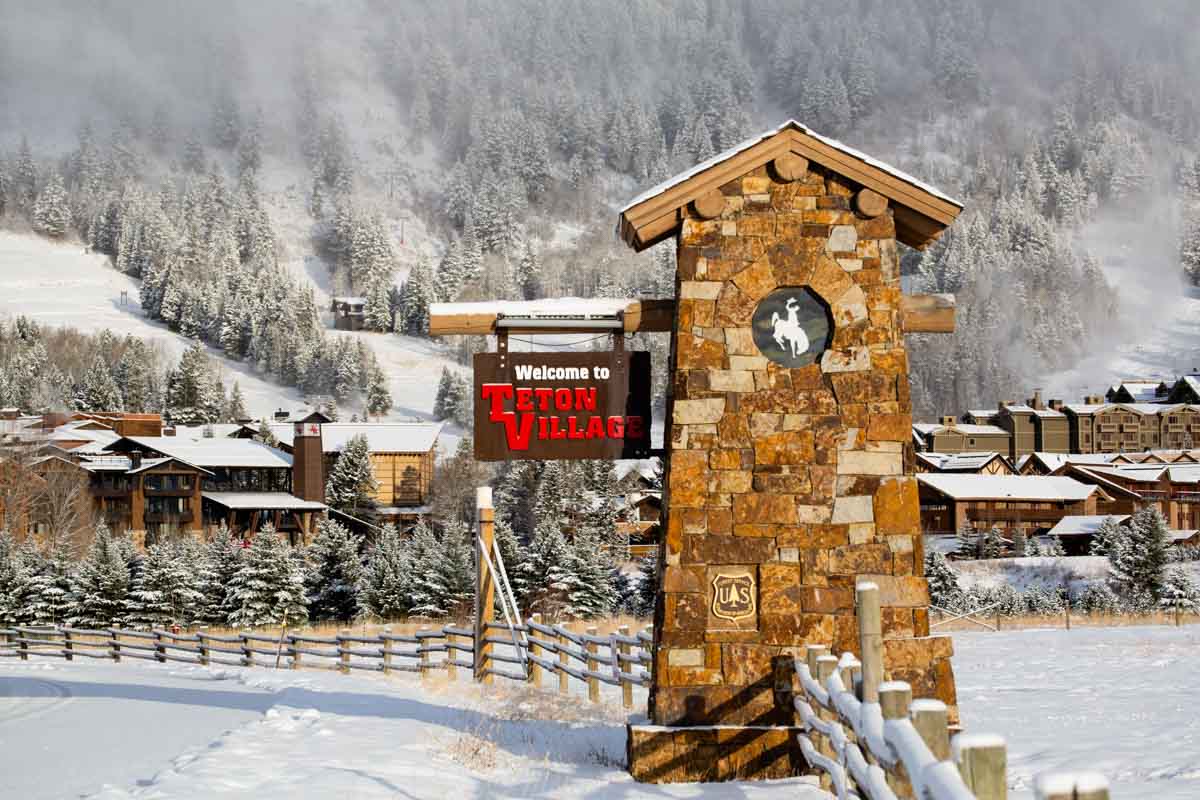 a stone pillar with Teton Village sign hanging from it, a snowy ski hill visible behind