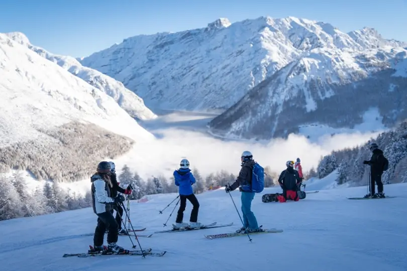 Skiers stand in the shadow on piste, looking towards a stellar view of high mountains peaking over a cloud-filled valley