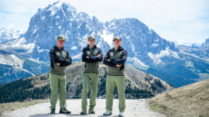 Three Dolomites Rangers standing on a mountain trail with the dramatic snow-capped peaks of Val Gardena in the background.