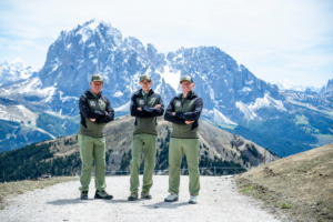 Three Dolomites Rangers standing on a mountain trail with the dramatic snow-capped peaks of Val Gardena in the background.