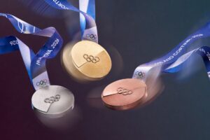 A dynamic image of the Olympic medals in motion, their textures catching the light. The iconic Milano Cortina 2026 ribbon swirls through the air, adding a sense of celebration and movement to the medal design.