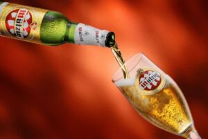 Bottle of Mutzig beer pouring into a branded glass with a foamy head, set against a warm red background.