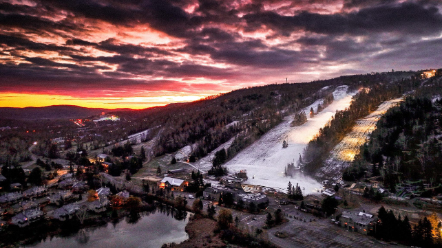 a sunset over white ski hills surrounded by brown trees