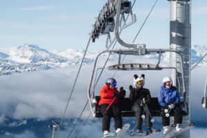 three ride a chairlift with the bar up, one wearing a panda helmet cover, who is waving at the camera