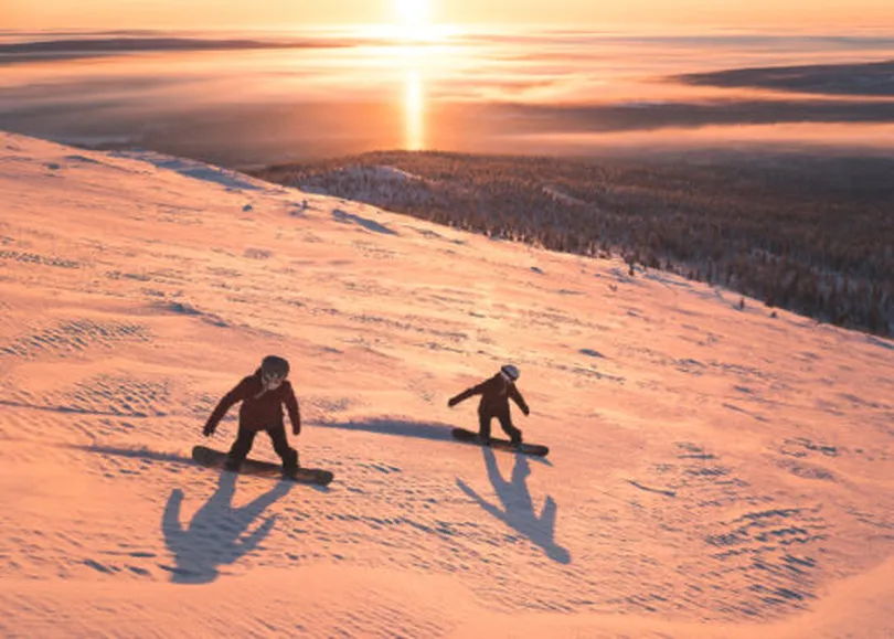 two snowboarders ride down an empty piste at dusk, an orange sunset lighting the slope