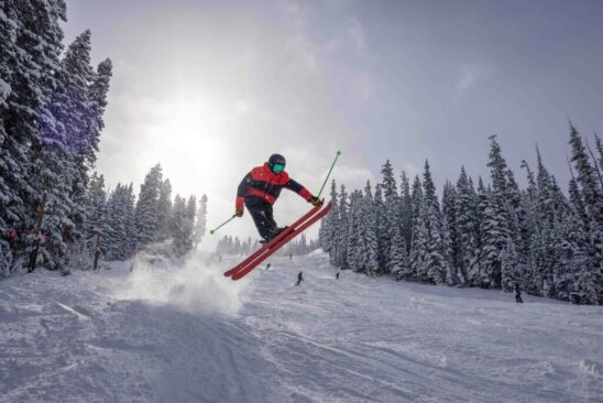 a skier takes air off a sidehit, by the piste, with tall snowy trees lining the side of the slope with several other skiers on