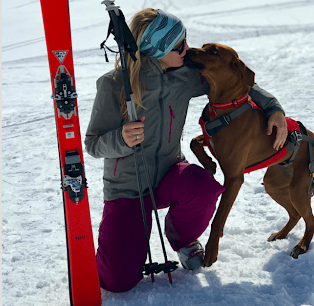 woman kisses ridgeback breed dog on the lips, kneeling on snow by red skis 