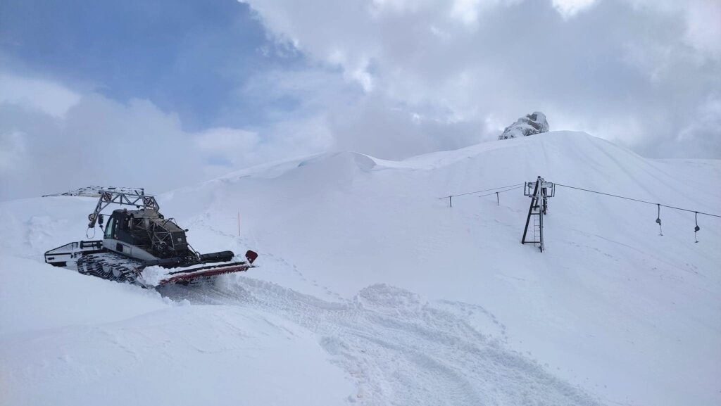 a piste basher works at the top of the mountain, a poma lift behind buried in snow