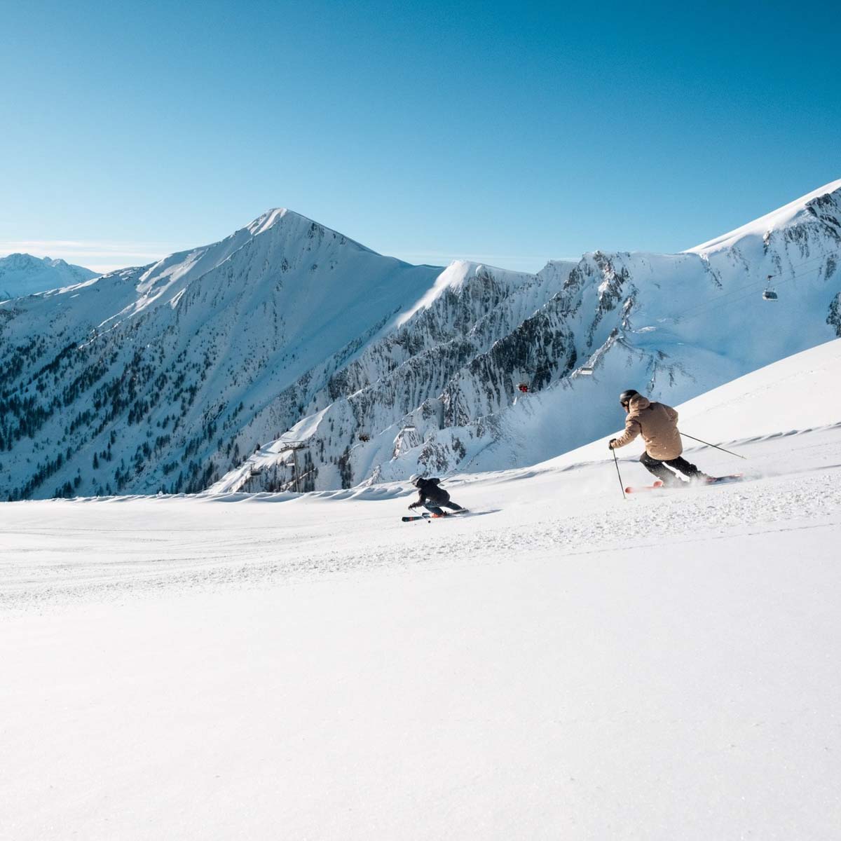 two skiers chase each other down a white piste, surrounded by white coated mountains