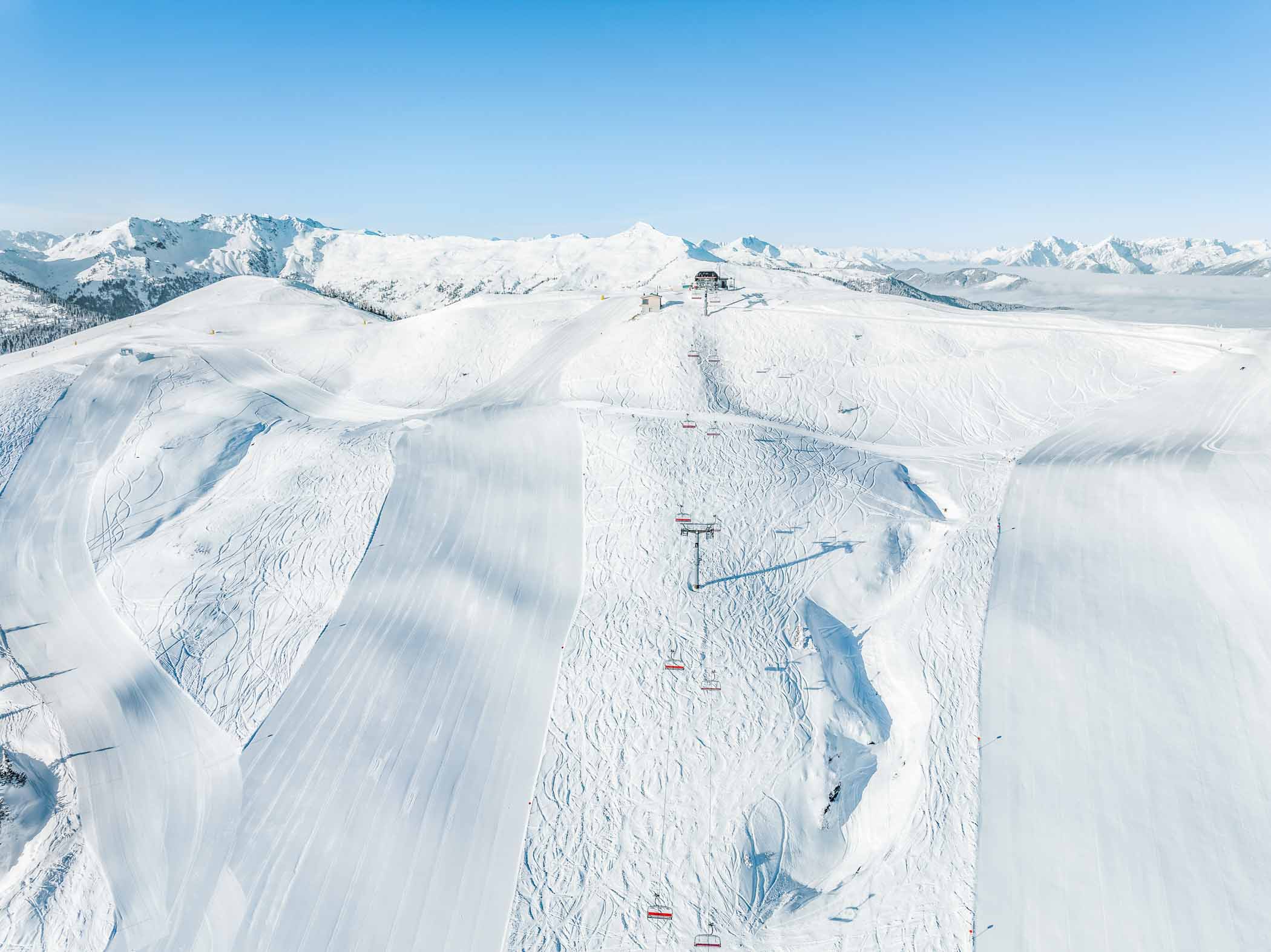 Beautiful white pistes, high in the mountains, with peaks in the distance poking through the clouds, a lift line running down the middle of the image (shot from above by drone)