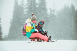 a mum and babe sit on a wooden sled in colourful ski kit in a very snowy environment