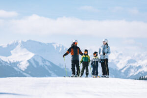 a family stand at the top of a mountain on a piste, posing for the camera - a big mountain vista behind on a sunny day