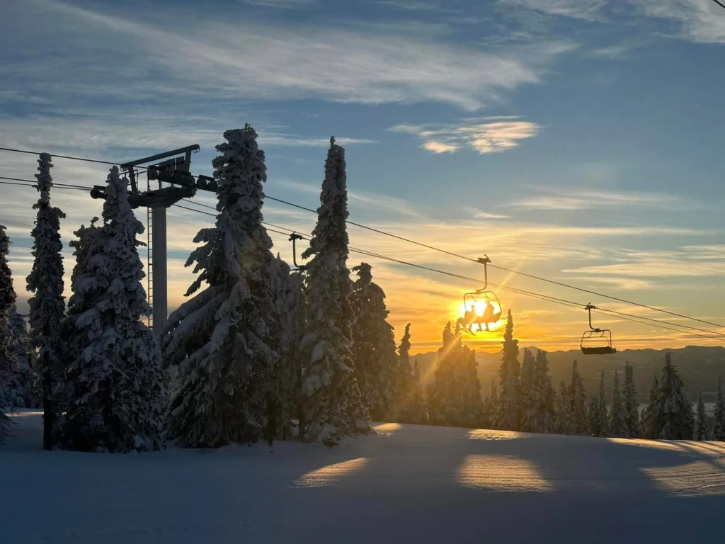 a chairlift loaded with skiers sits silhouetted right in front of the sun, huge trees covered in snow shaded