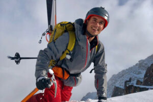 man with skis attached to pack is on knee and hands as he, smiling big, climbs over the top ledge of a snowy mountain