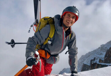 man with skis attached to pack is on knee and hands as he, smiling big, climbs over the top ledge of a snowy mountain