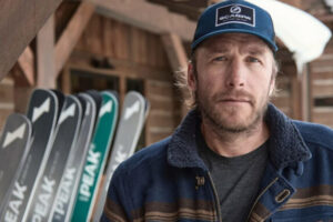 Bode Miller looks seriously into the camera, some skis stacked behind, tips just visible but blurred out