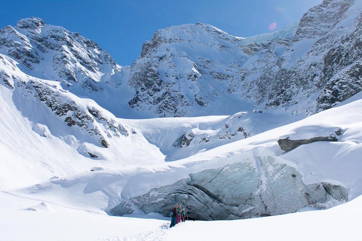 a line of ski tourers enter a glacier cave below big snow-covered and steep mountains under a blue sky