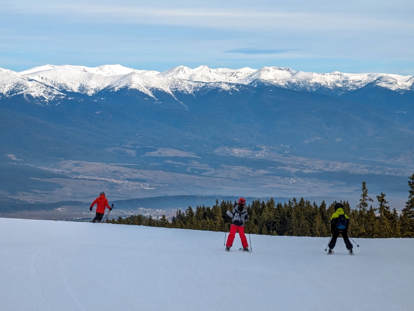 two kids skiing on the shade, a snowless valley below with just mountain tops white capped