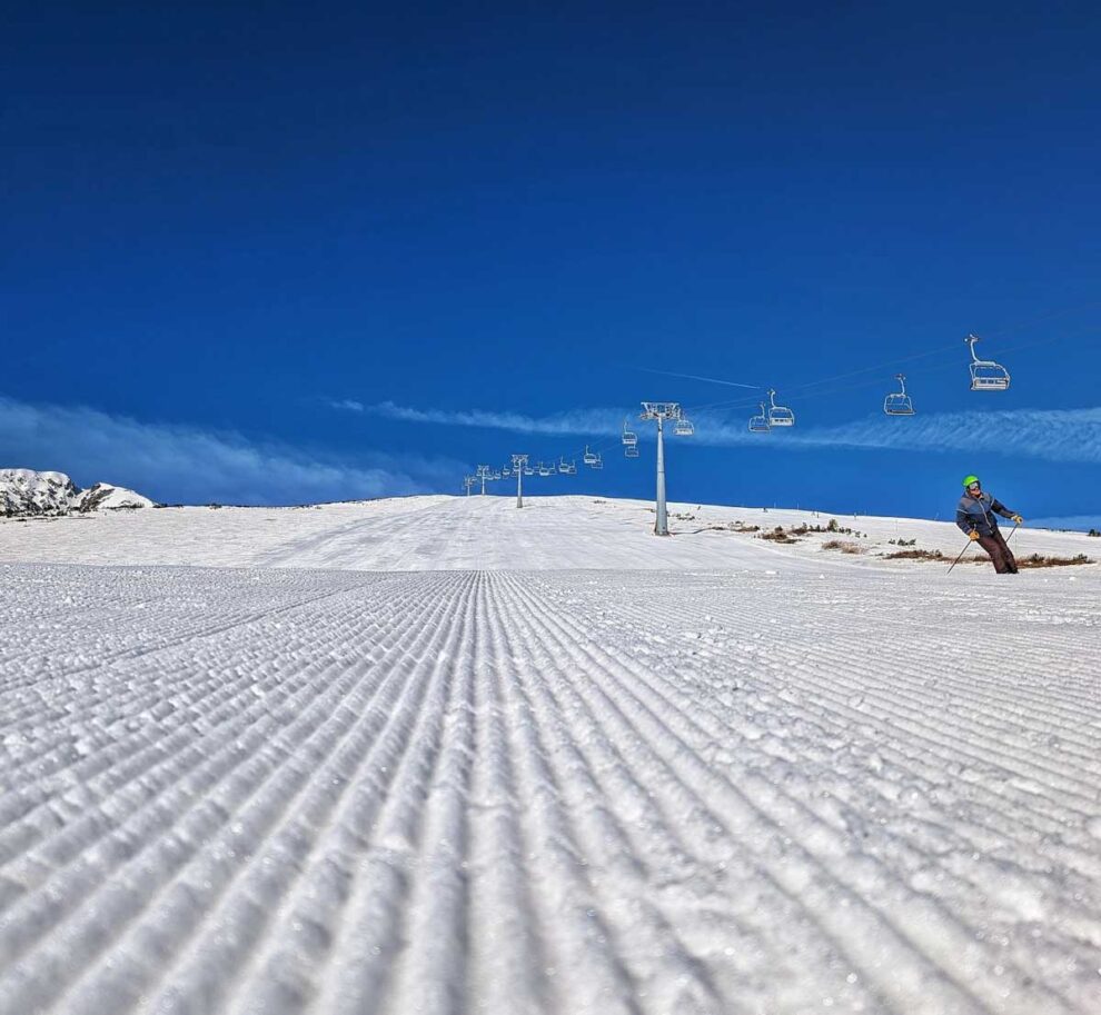 crystally piste corduroy is pictured on a bluebird day, an empty chair overhead and a lone skier descending the groomer