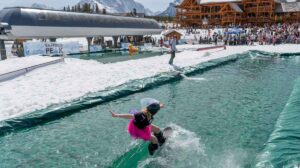 woman snowboarder in pink tutu pond skims towards a big crowd, high in the mountains judging on the peaks in background