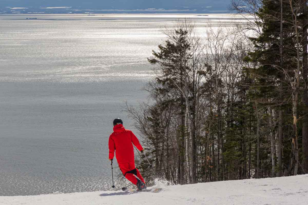 red telemark skier skis down towards a huge body of shimmering water that is a lake below