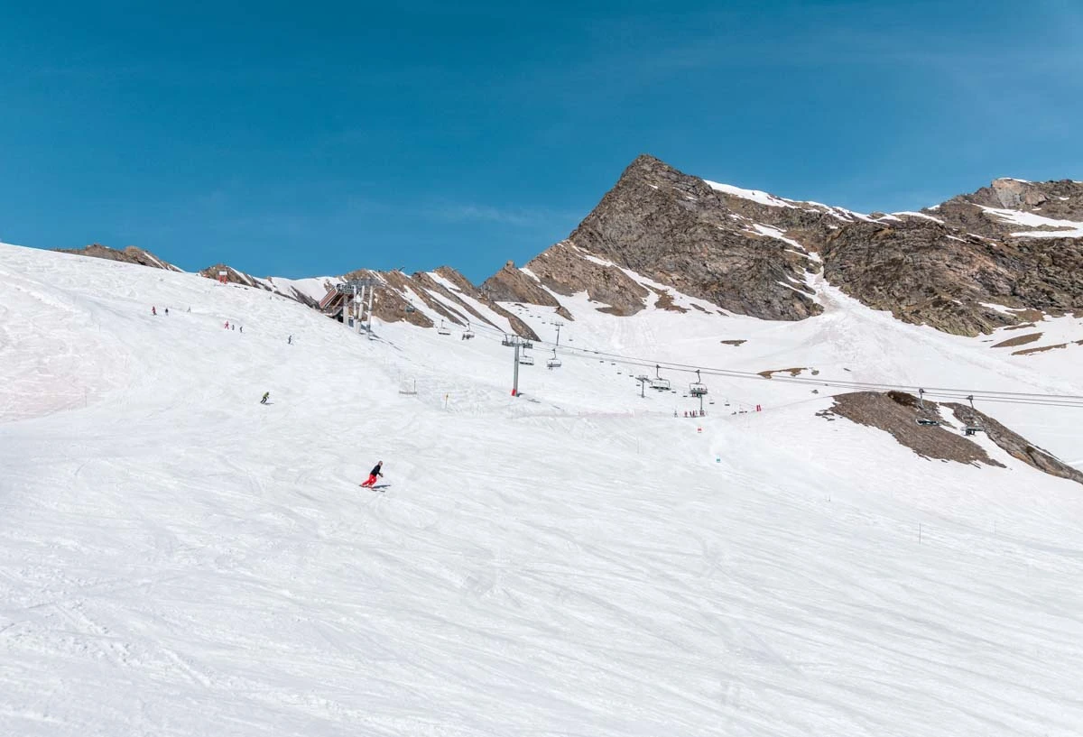 a top-of-mountain scene with two chairlifts serving gentle pistes and a few skiers enjoying the snow