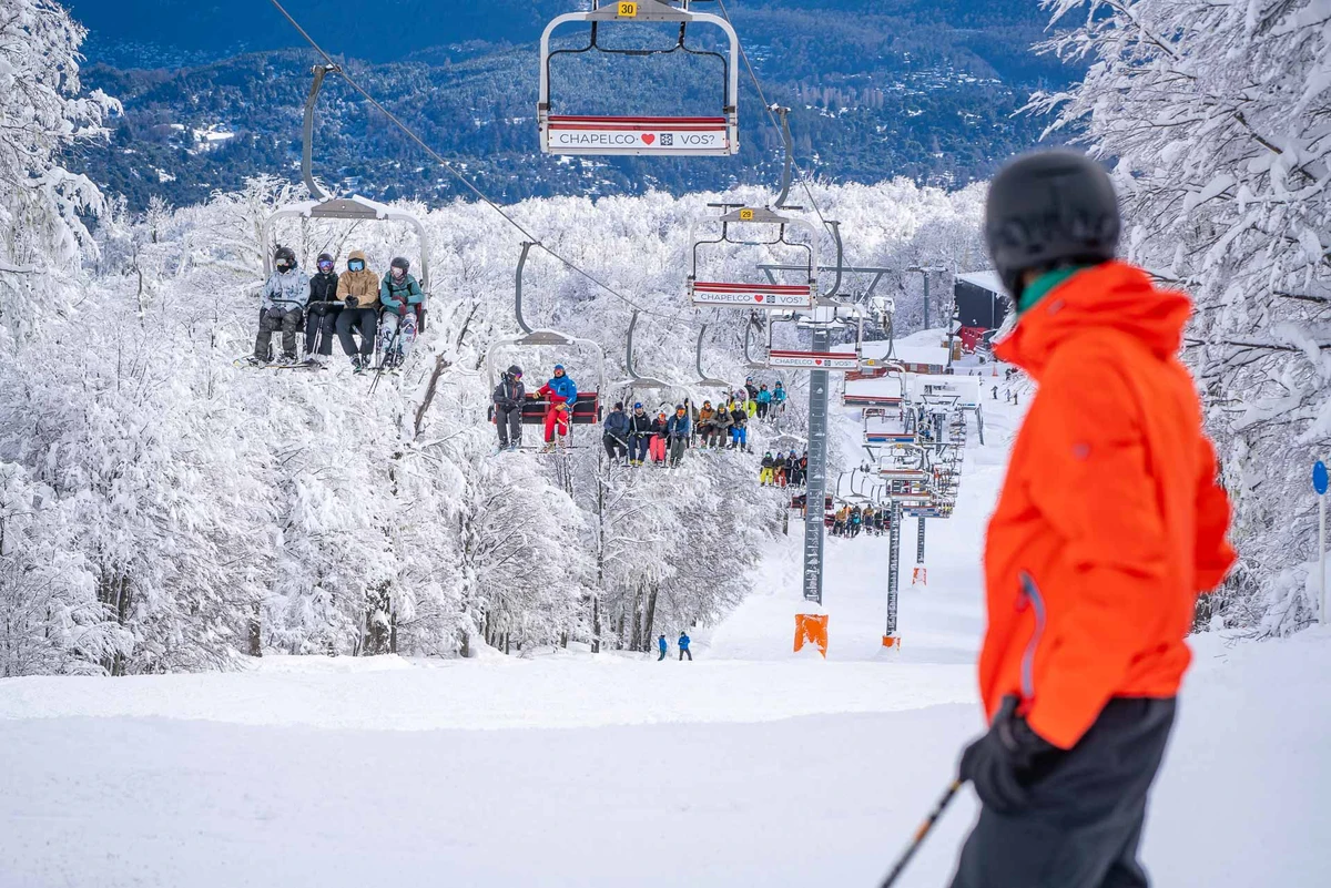 a blurred skier in orange jacket dominates the foreground, looking over a packed chairlift travelling over a freshly snowed-on piste through trees covered in snow