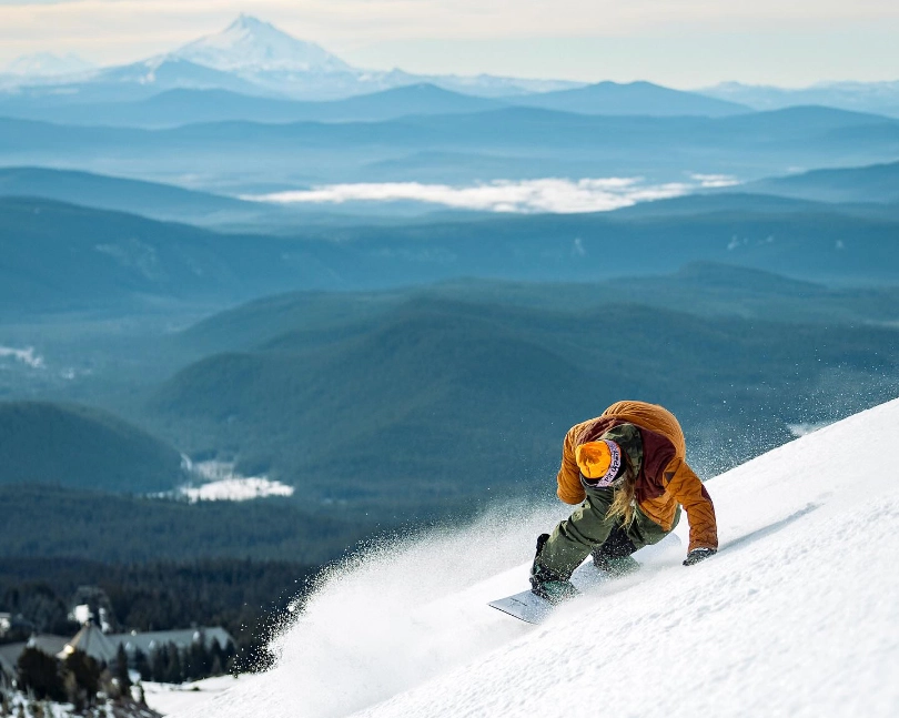 a snowboarder on his toe-edge, looks down the steep hill, the valley and mountains beyond/below beautiful and blue in silhouette, a snowy peak visible in long distance