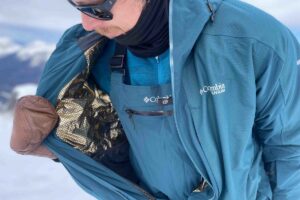 skier on slope opens jacket to show the gold interior of the ski kit, wearing leather mittens and sunglasses, with bibbed ski pants beneath jacket