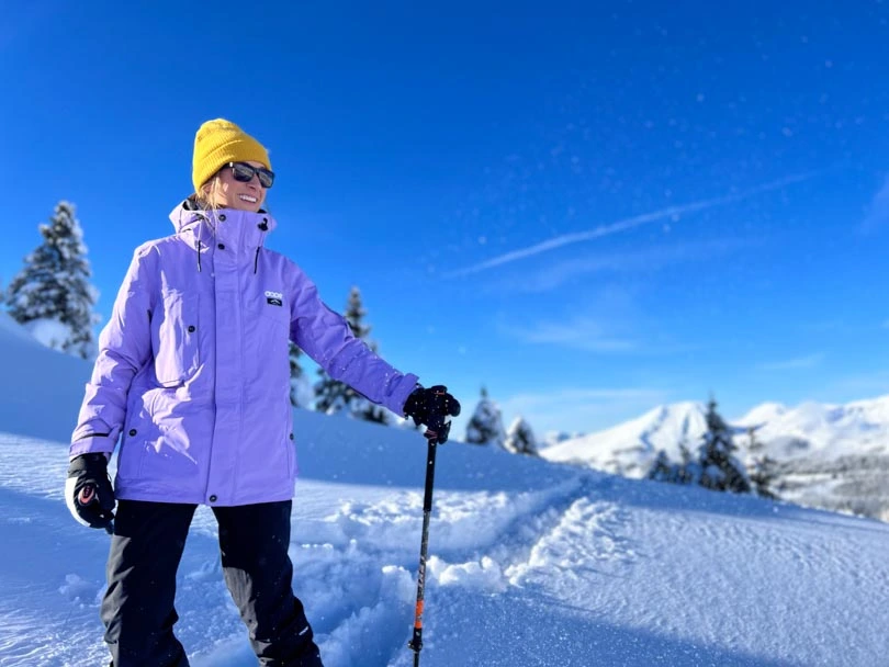 Skier in pastel purple ski jacket and yellow hat, smiles beyond camera on a bluebird day on snow