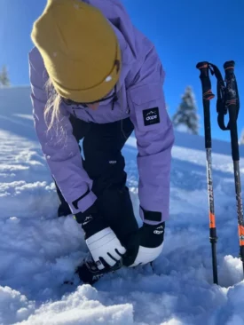 a skier bends down to buckle ski boot, wearing white and black leather gloves, a purple jacket and a yellow hat, standing on snow
