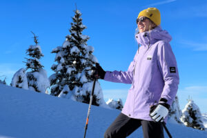 Skier in pastel purple ski jacket and yellow hat, smiles beyond camera on a bluebird day on snow