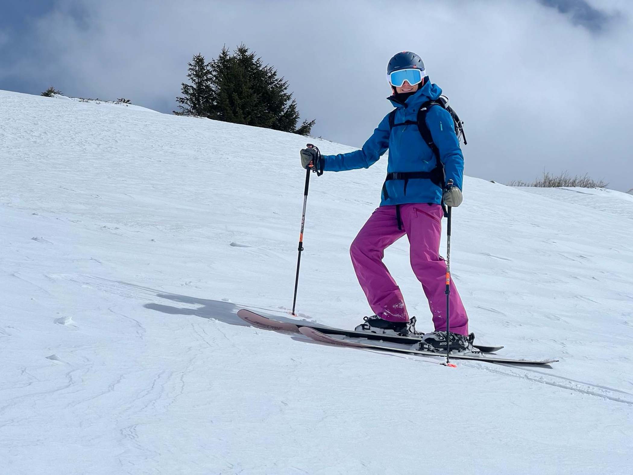 skier in purple ski pants and blue jacket, stands on the slope smiling at camera, with goggles on