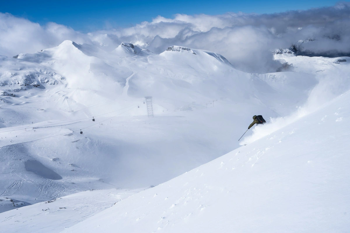 a skier alone off-piste, skis fast down a white snowy face, the gondola and ski area visible in the distance below