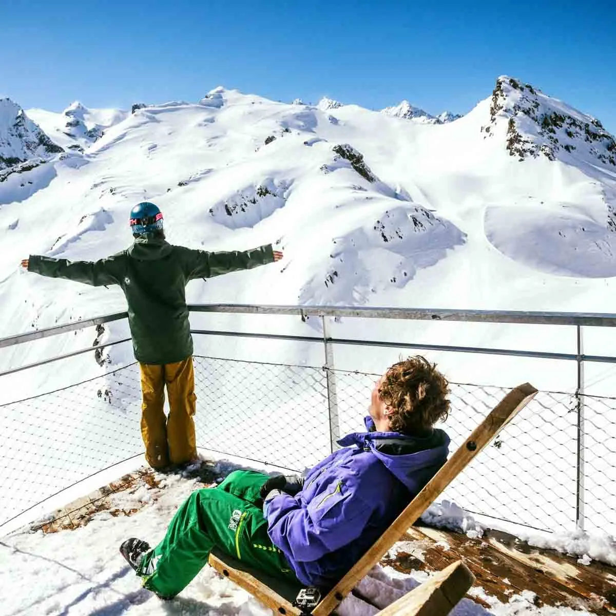 one man in deck chair, another arms open wide on an elevated mountain terrace looking over the top of a ski area