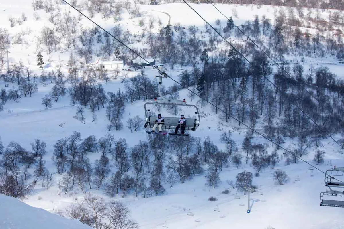 a chairlift with two snowboarders travels above snowy slopes and bare trees