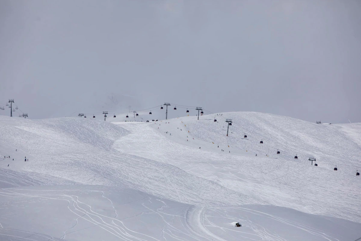 a high section of a ski area, above the treeline, is photographed, with a lift and a couple of slopes with ant-sized skiers