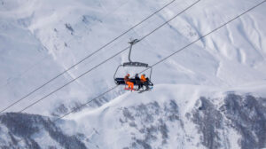 three snowboarders on a chairlift, looking small against a huge mountain behind