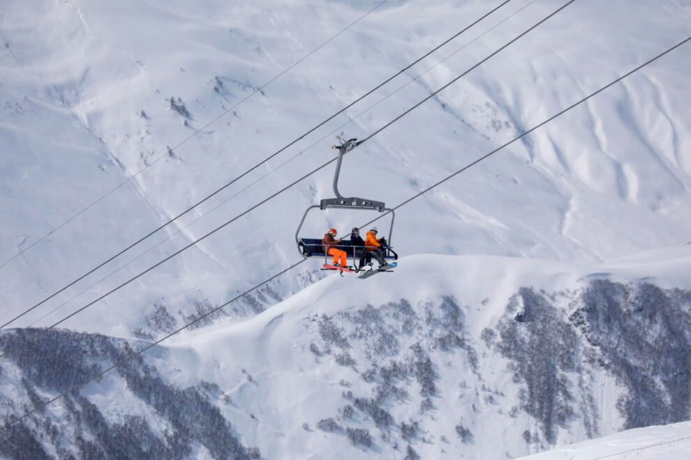 three snowboarders on a chairlift, looking small against a huge mountain behind