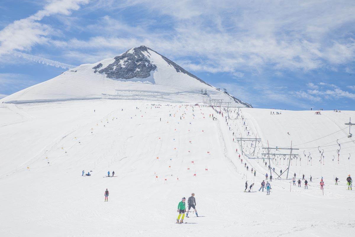 a glacier ski area photographed from the bottom of the gentle, low-angled slope, with a peak at the top of the slope, rising above the area, and a t-bar line to the right of the image, with skiers riding it