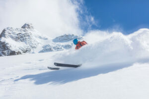 blue sky, fresh snow, mountain tops, and a skier in orange skiing in powder