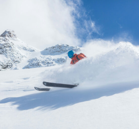 blue sky, fresh snow, mountain tops, and a skier in orange skiing in powder