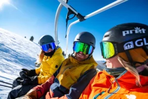 three friends laugh on a chairlift, all wearing reflective lens ski goggles, the focus of the photo